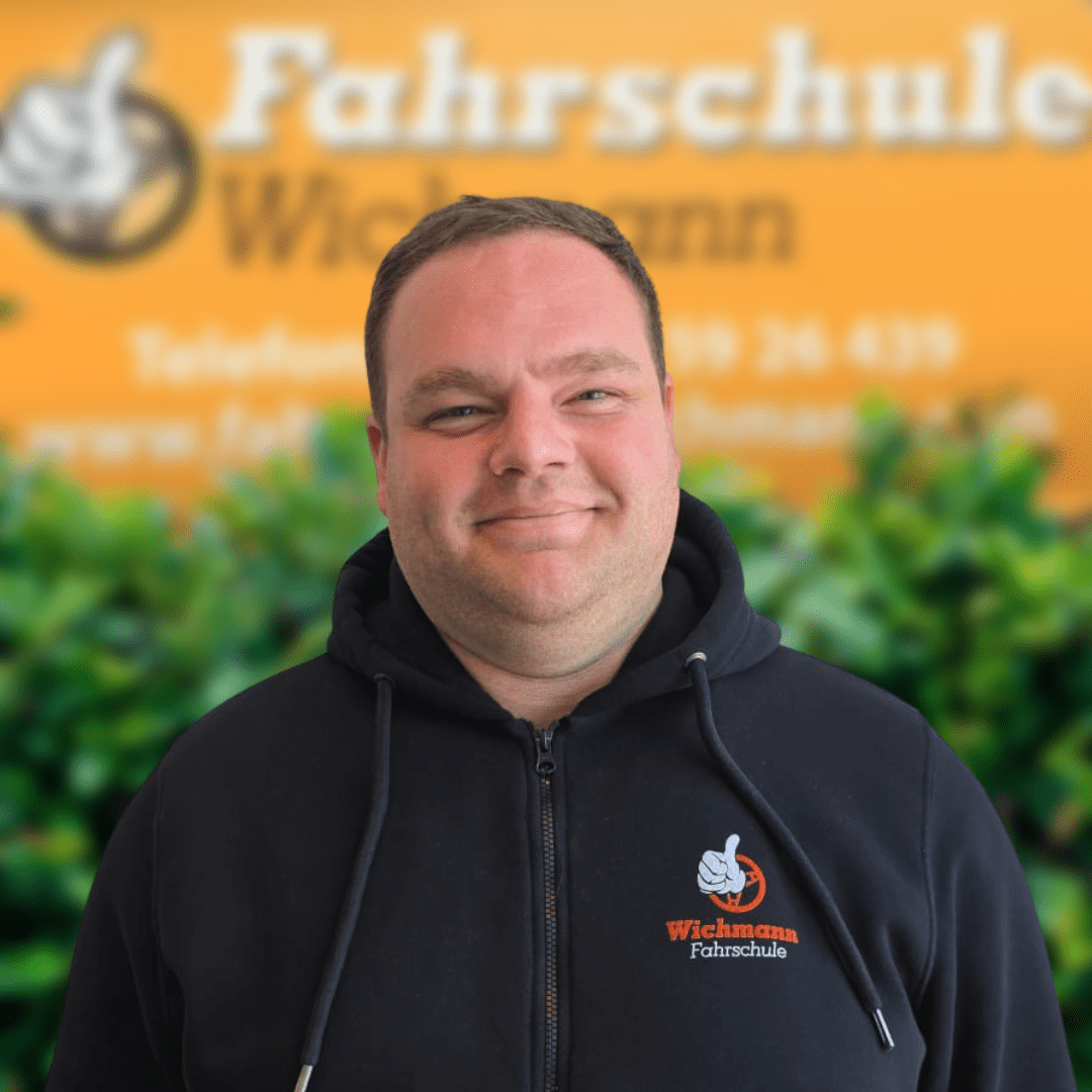 KilianFahrlehrerFahrschuleWichmann Fahrschule Wichmann Ganderkesee Autoführerschein LKW Führerschein Motorradführerschein Gunnar Fahrlehrer der Fahrschule Wichmann Delmenhorst lächelt vor dem gelben Firmenschild mit Logo und T Shirt