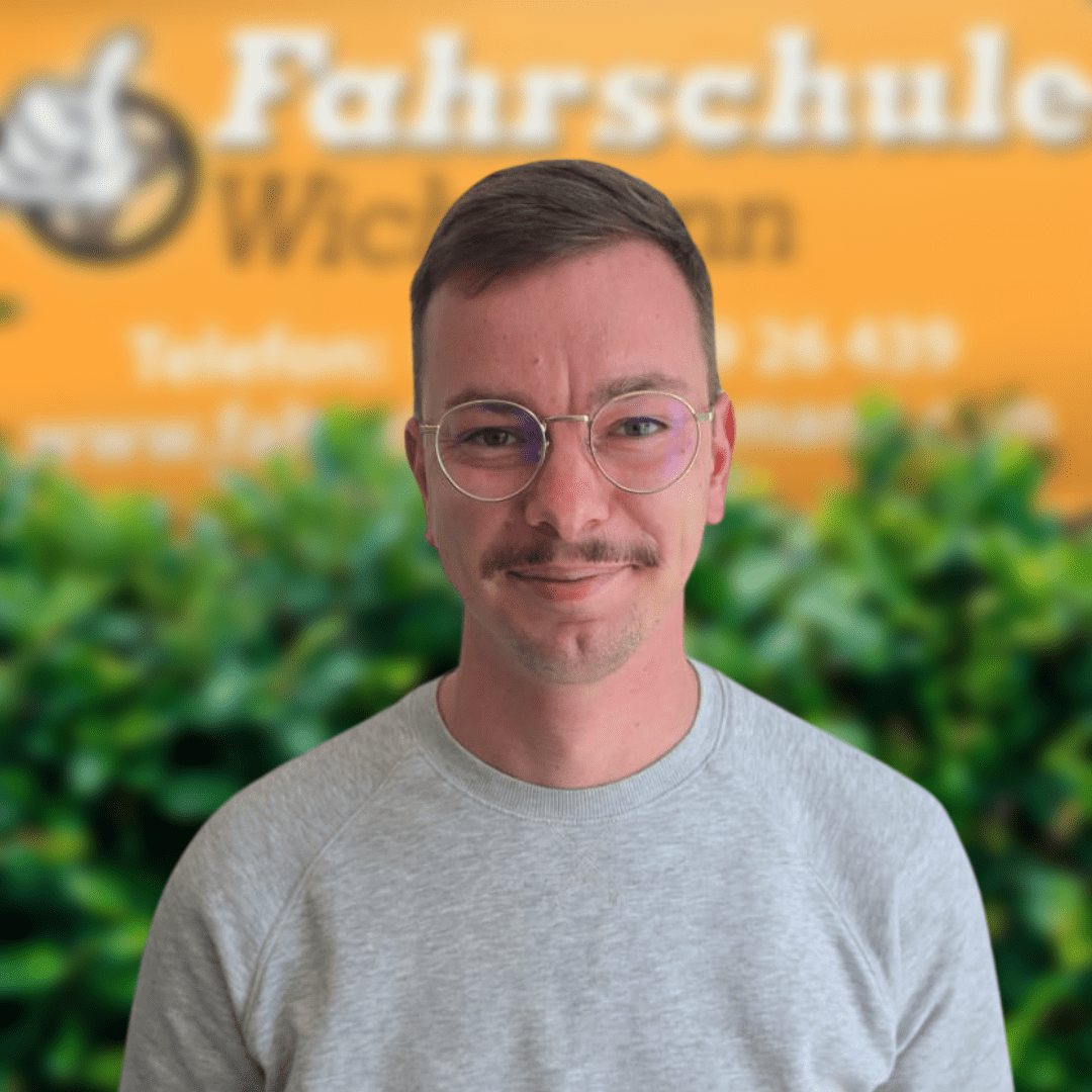 TillFahrlehrerFahrschuleWichmann Fahrschule Wichmann Ganderkesee Autoführerschein LKW Führerschein Motorradführerschein Fahrlehrer Sofiane der Fahrschule Wichmann Delmenhorst vor grünem Busch und gelbem Firmenschild